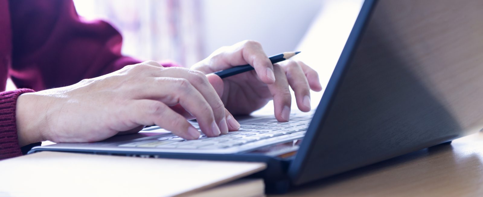 Hands of a man in red sweater holding a pencil while typing on laptop computer notebook keyboard in the office.