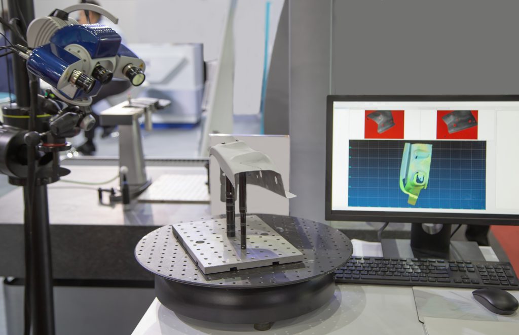 An optical 3D measurement instrument pointed at a metal part, with a digital model of the part on a laptop screen.