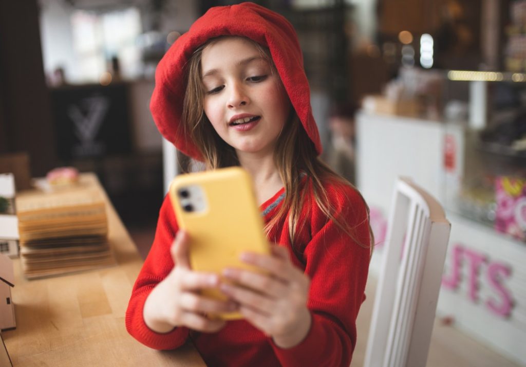 A young girl in a red hoodie holding a yellow mobile phone.